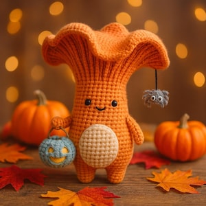 May include: A handmade orange crochet mushroom figure with a smiling face, holding a blue pumpkin-shaped treat bag. A small gray spider hangs from the mushroom's cap. Pumpkins and fall leaves are in the background.