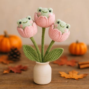 May include: A handcrafted crochet bouquet featuring three smiling frog figures nestled in pink tulip-shaped flowers. The arrangement includes green stems and leaves, presented in a small white vase. The background includes pumpkins and autumn leaves.