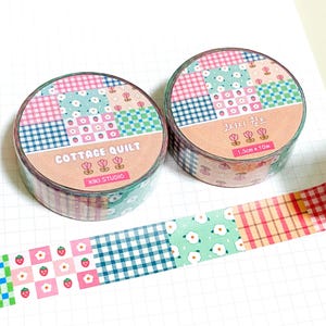 Pode incluir: Dois rolos de fita adesiva decorativa com um design de colcha de cabana. A fita apresenta padrões de patchwork rosa, azul e verde com motivos florais e de morango. O texto "COTTAGE QUILT" está impresso no rótulo.