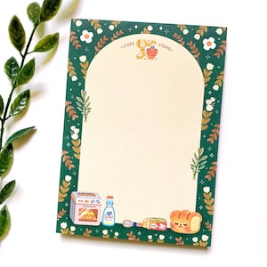 May include: A notepad with a green border featuring floral and leaf designs. The notepad has a cream-colored interior with the words ".cozy vibes." at the top. The bottom of the notepad has illustrations of baking supplies, including an oven, a bottle, a whisk, butter, and bread.