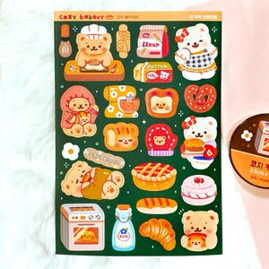 Op de afbeelding: Een vel stickers met een gezellig bakkerijthema. De stickers bevatten schattige berenpersonages en diverse bakkerijproducten zoals brood, croissants en taarten. Het kleurenpalet omvat tinten bruin, wit en groen. Tekst op het vel luidt "COZY BAKERY".