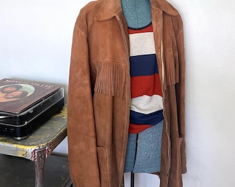 May include: A brown suede jacket with fringe detailing on the chest and sleeves. The jacket is displayed on a mannequin, revealing a striped red, white, and blue shirt underneath. A vintage record player sits on a nearby shelf.