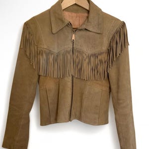 May include: A tan suede jacket with fringe detailing across the chest and shoulders. The jacket has a collared neckline, a zipper closure, and long sleeves. The jacket is hanging on a wooden hanger.