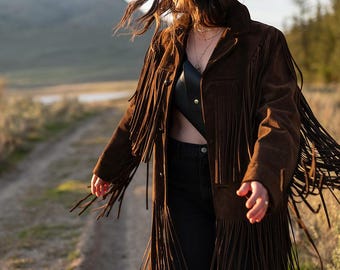 May include: A brown suede fringe jacket with long sleeves and a button-down front. The jacket is worn over a black top and black jeans. The image is taken outdoors on a dirt path.