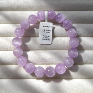 Puede incluir: Una pulsera circular hecha de cuentas de kunzita natural de color morado claro pulidas. Se adjunta una etiqueta rectangular blanca con el texto E084210, 11.3mm/6.75in y Natural Kunzite Bracelet. Las cuentas tienen un acabado liso y translúcido.