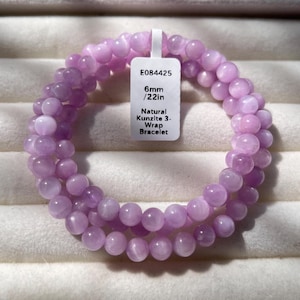 May include: A triple-wrap bracelet made of round, light purple natural kunzite beads. A white rectangular tag is attached, featuring the text E084425, 6mm / 22in, and Natural Kunzite 3-Wrap Bracelet. The beads have a smooth, polished finish.