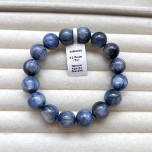 May include: A stretch bracelet composed of round, polished natural kyanite beads in shades of blue with white streaks. A white tag attached to the bracelet displays the text E084430, 12.8mm / 7in, and Natural Kyanite Bracelet in black print.