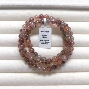 May include: A multi-strand bracelet composed of 6mm round beads in shades of black, gold, and reddish-brown. A white rectangular tag is attached, displaying the text E084258, 6mm / 20in, and Black Gold Super Seven Bracelet in black lettering.