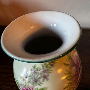 May include: A small, round ceramic vase with a wide, flared rim. The vase is decorated with a floral pattern featuring purple and pink flowers with green leaves on a pale yellow background. The rim has a green trim.