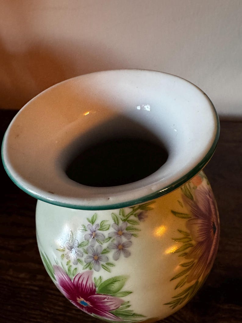 May include: A small, round ceramic vase with a wide, flared rim. The vase is decorated with a floral pattern featuring purple and pink flowers with green leaves on a pale yellow background. The rim has a green trim.