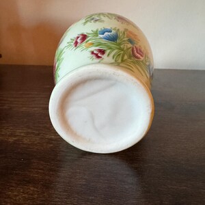 May include: A ceramic vase with a light green background and a floral pattern featuring red, blue, and purple flowers. The vase has a wide, white base and a rounded body. The flowers are surrounded by green leaves.