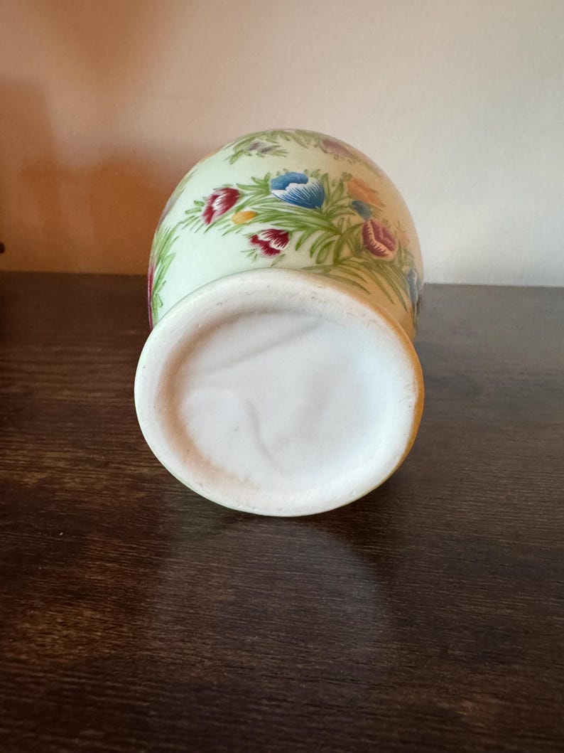 May include: A ceramic vase with a light green background and a floral pattern featuring red, blue, and purple flowers. The vase has a wide, white base and a rounded body. The flowers are surrounded by green leaves.