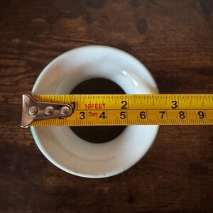 May include: A white ceramic ring with a green rim is measured with a yellow tape measure. The tape measure shows the ring is approximately 2 inches in diameter. The ring sits on a dark brown wooden surface.