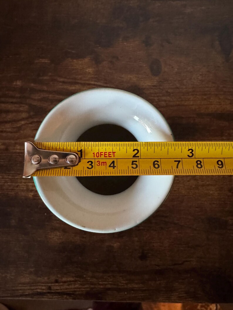 May include: A white ceramic ring with a green rim is measured with a yellow tape measure. The tape measure shows the ring is approximately 2 inches in diameter. The ring sits on a dark brown wooden surface.