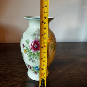 May include: A light green ceramic vase with floral designs in pink, red, blue, and yellow. The vase is approximately 8 inches tall. The vase has a gold rim and a white base. A measuring tape is next to the vase.
