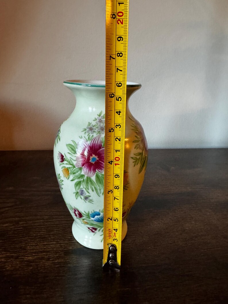May include: A light green ceramic vase with floral designs in pink, red, blue, and yellow. The vase is approximately 8 inches tall. The vase has a gold rim and a white base. A measuring tape is next to the vase.