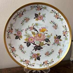 May include: A white ceramic plate with a gold rim and floral design. The plate features a central arrangement of pink, yellow, and blue flowers with dark green leaves. Smaller floral clusters encircle the plate's edge, creating a decorative and elegant aesthetic.