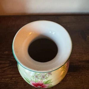May include: A small, round ceramic vase with a wide opening. The vase features a floral design with pink and white flowers on a pale yellow background. The rim is white with a green trim.