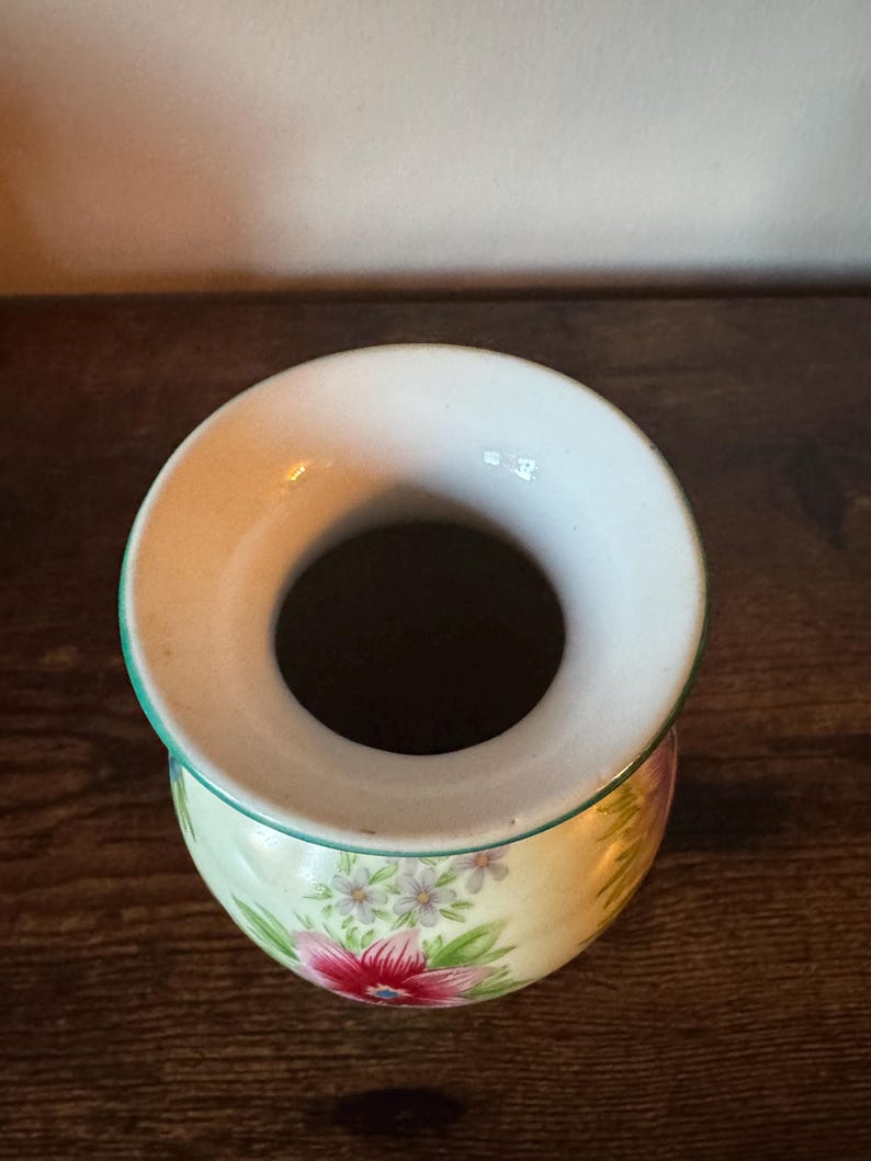May include: A small, round ceramic vase with a wide opening. The vase features a floral design with pink and white flowers on a pale yellow background. The rim is white with a green trim.