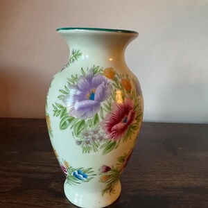 May include: A cream-colored ceramic vase with a green rim and a flared top. The vase is adorned with a floral pattern featuring purple, red, and yellow flowers with green leaves. The vase has a rounded base.