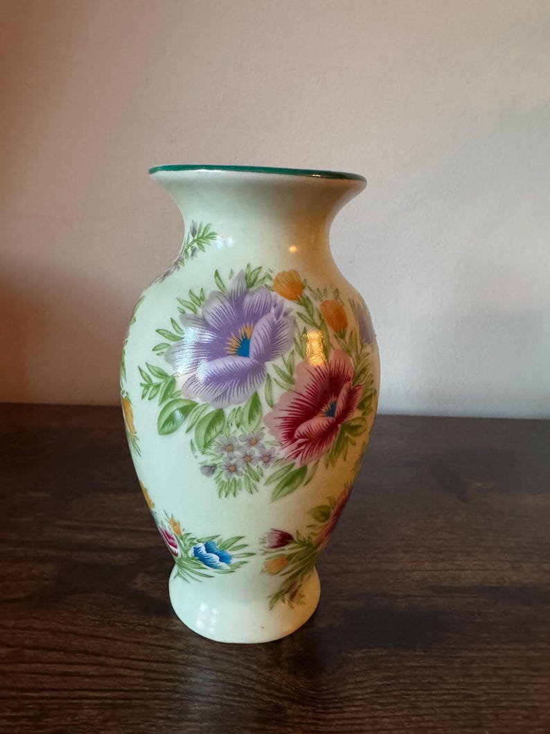 May include: A cream-colored ceramic vase with a green rim and a flared top. The vase is adorned with a floral pattern featuring purple, red, and yellow flowers with green leaves. The vase has a rounded base.