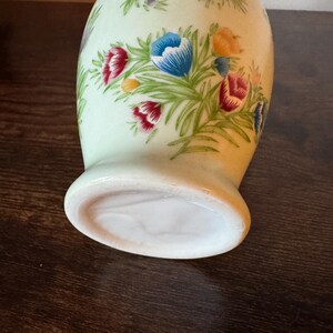 May include: A ceramic vase with a light green background and a floral design. The flowers are in shades of red, blue, purple, and yellow, with green leaves. The vase has a rounded body and a flared base.