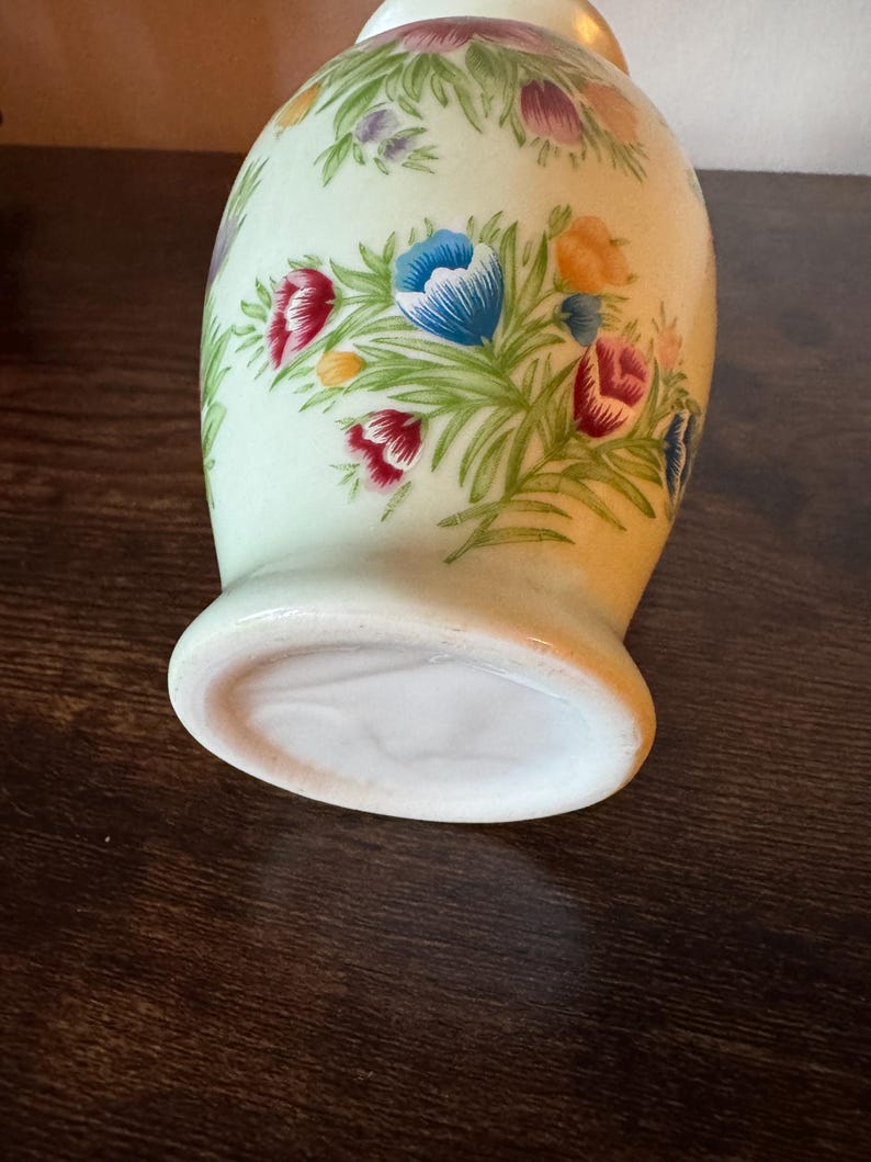 May include: A ceramic vase with a light green background and a floral design. The flowers are in shades of red, blue, purple, and yellow, with green leaves. The vase has a rounded body and a flared base.