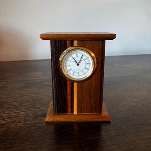 May include: A wooden desk clock with a gold-colored bezel and a white clock face with black Roman numerals. The clock is set in a rectangular wooden frame with a multi-toned wood grain design. The clock measures approximately 5 inches tall.
