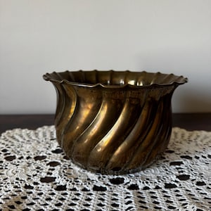 May include: A bronze-colored, antique-style planter with a fluted, spiral design. The rim has a scalloped edge. The planter sits on a white, crocheted doily. The background is a neutral color.