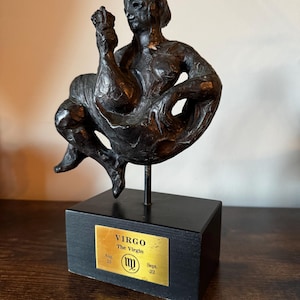 May include: A bronze sculpture of a seated figure, holding an object aloft. The sculpture is mounted on a black rectangular base with a gold plaque that reads "VIRGO The Virgin". The plaque also includes the Virgo symbol and dates.