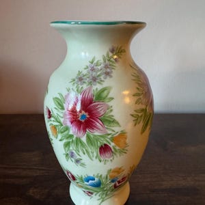 May include: A light green ceramic vase with a flared top and a round base. The vase is decorated with a floral pattern featuring red, pink, blue, and yellow flowers with green leaves. The rim is trimmed with a teal color.