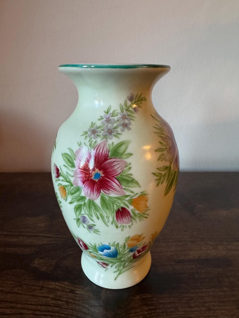 May include: A light green ceramic vase with a flared top and a round base. The vase is decorated with a floral pattern featuring red, pink, blue, and yellow flowers with green leaves. The rim is trimmed with a teal color.