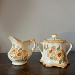 May include: Cream-colored ceramic creamer and sugar bowl set. The set features a floral design with yellow and brown flowers and green leaves. The sugar bowl has a lid and two handles. The creamer has a handle and a spout.