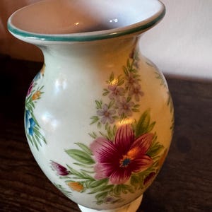 May include: A cream-colored ceramic vase with a flared top and a white base. The vase is adorned with a floral pattern featuring pink, purple, blue, and orange flowers with green leaves. The rim is trimmed with a green border.