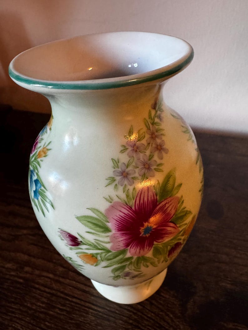 May include: A cream-colored ceramic vase with a flared top and a white base. The vase is adorned with a floral pattern featuring pink, purple, blue, and orange flowers with green leaves. The rim is trimmed with a green border.