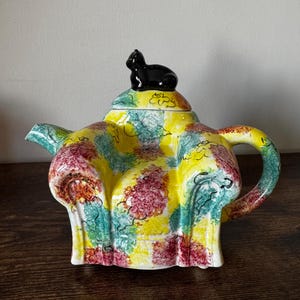 May include: A ceramic teapot with a black cat figurine on the lid. The teapot features a yellow base with colorful floral patterns in red, green, and blue. The handle and spout are also decorated with the same pattern.