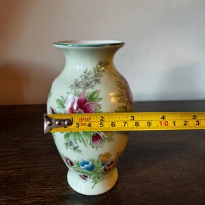May include: A small, light green ceramic vase with floral designs in red, blue, and yellow. The vase is approximately 4 inches tall and has a green rim. A measuring tape is placed across the vase.