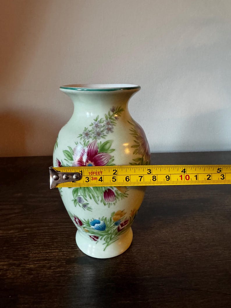 May include: A small, light green ceramic vase with floral designs in red, blue, and yellow. The vase is approximately 4 inches tall and has a green rim. A measuring tape is placed across the vase.