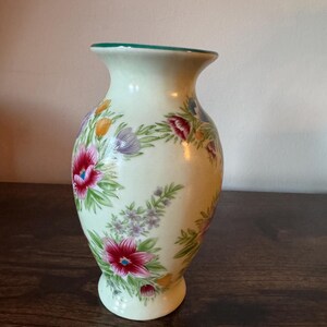 May include: A cream-colored ceramic vase with a flared top and a round base. The vase is decorated with a floral pattern featuring red, purple, and yellow flowers with green leaves. The rim of the vase is painted green.