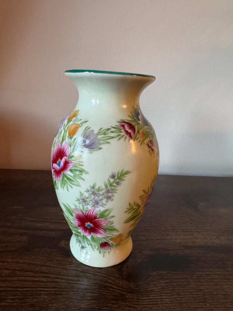 May include: A cream-colored ceramic vase with a flared top and a round base. The vase is decorated with a floral pattern featuring red, purple, and yellow flowers with green leaves. The rim of the vase is painted green.