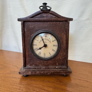 May include: A dark brown wooden mantel clock with a triangular top and a metal ring handle. The circular clock face features black Roman numerals and the text Colonial Clock Co. estb. 1870. The clock stands on four small, rounded feet.