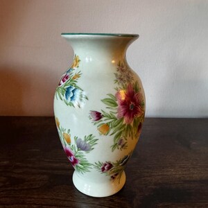 May include: A cream-colored ceramic vase with a flared top and a round base. The vase is adorned with colorful floral patterns in shades of pink, blue, yellow, and purple, with green leaves. The rim has a green trim.