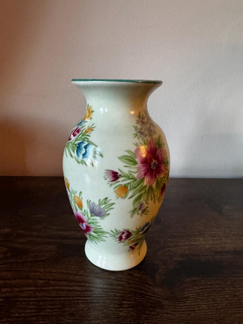 May include: A cream-colored ceramic vase with a flared top and a round base. The vase is adorned with colorful floral patterns in shades of pink, blue, yellow, and purple, with green leaves. The rim has a green trim.