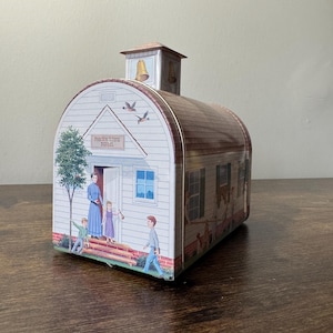 May include: A decorative tin shaped like a schoolhouse, featuring a bell tower and a brown roof. The front of the tin displays the words "Prairie Town School" above a doorway with figures of people. The sides depict children and other scenes.