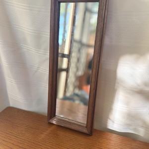 May include: A rectangular mirror featuring a dark brown wooden frame with a beveled edge. The mirror glass reflects a bright outdoor scene, and the frame has a smooth, polished finish with visible wood grain patterns throughout the structure.