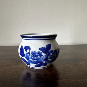 May include: A small, round white ceramic pot with a blue floral design. The pot features a band of blue around the rim and a dark blue floral pattern of roses and leaves. The pot is sitting on a dark brown wooden surface.
