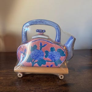 May include: A ceramic teapot with a pink body, blue floral design, and blue handle and spout. The teapot sits on a small wooden stand. The teapot is wrapped in clear plastic.
