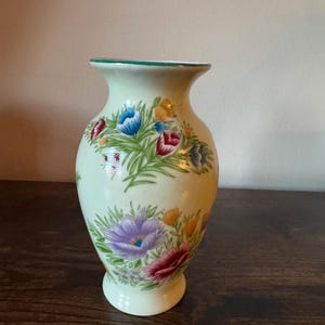 May include: A light green ceramic vase with a flared top and a round base. The vase is decorated with colorful floral patterns in red, blue, purple, and yellow. The rim of the vase has a green trim.