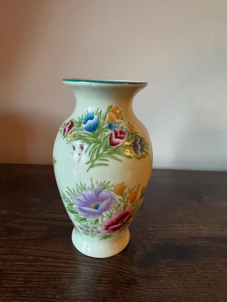 May include: A light green ceramic vase with a flared top and a round base. The vase is decorated with colorful floral patterns in red, blue, purple, and yellow. The rim of the vase has a green trim.