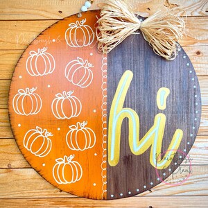 May include: Round wooden fall-themed sign with a two-tone design. The left side is orange with white pumpkin outlines, and the right side is brown with the word "hi" in yellow. A raffia bow and beaded string add detail.